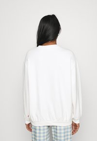 Sudadera blanca de ajuste relajado, con puños y dobladillo acanalados. Presenta un cuello redondo y hombros caídos. Llevada con pantalones de cuadros azules.