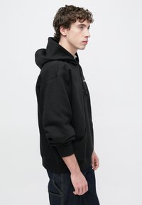 Sudadera negra con capucha, bolsillo frontal, cordón ajustable y puños acanalados. Material suave, corte relajado, costuras visibles y diseño minimalista.