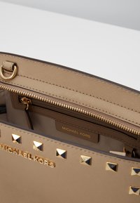 Beżowa torebka z złotym wykończeniem, z teksturowaną powierzchnią, zamykaną na zamek kieszonką oraz piramidkowymi ćwiekami wzdłuż górnej krawędzi. Widoczna marka.