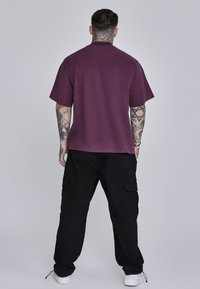 Camisa corta de manga en color burdeos con un corte relajado, combinada con pantalones cargo negros que cuentan con bolsillos laterales y zapatillas blancas. Diseño simple.