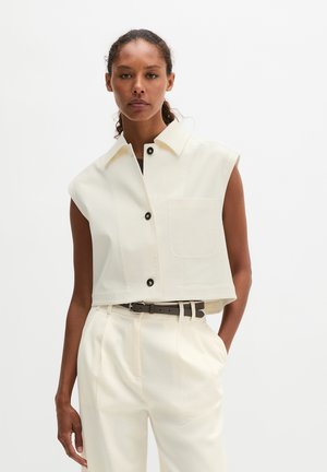 Femme portant une veste blanche courte sans manches avec des boutons noirs et un pantalon taille haute assorti, debout avec une main dans la poche.