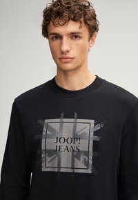 Felpa nera in cotone con collo a giro. Presenta un motivo grafico grigio con la Bandiera Britannica e il testo "JOOP! JEANS" al centro.