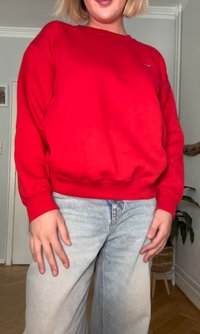 Red fleece sweatshirt with ribbed cuffs and hem, paired with light blue wide-leg jeans. Indoor setting with a plant in the background.