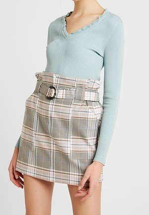 Femme portant un pull côtelé bleu clair à col en V et une mini-jupe à carreaux taille haute ceinturée avec poches devant.