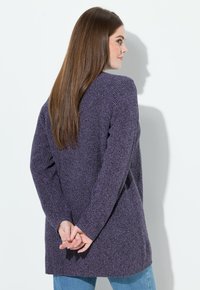 Kobieta o długich brązowych włosach, ubrana w fioletowy sweter z dzianiny i niebieskie dżinsy, stojąca z rękami splecionymi za plecami.