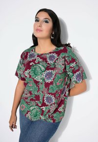 Burgunderfarbendes Blusenshirt mit grünen und blauen Paisleymustern, kurzen Ärmeln und rundem Halsausschnitt, kombiniert mit blauen Jeans. Glatte Stofftextur.