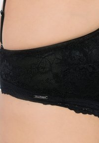 Černá krajková podprsenka s vlnitými okraji, hladkým povrchem a jemným květinovým vzorem. Má malou kovovou značku na levé straně.