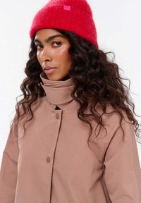 Kobieta z długimi kręconymi włosami nosi jasnoczerwoną dzianą czapkę i beżowy płaszcz z wysokim kołnierzem na jednolitym tle.