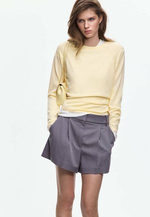 Joven mujer con un suéter amarillo pálido, pantalones cortos grises plisados y llevando un pequeño bolso amarillo colgado del hombro, de pie con las manos en los bolsillos.