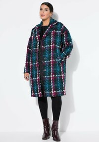 Geruite jas in teal, bordeaux en zwart met witte accenten, voorzien van een gelobde kraag, knoopsluiting en oversized silhouet.
