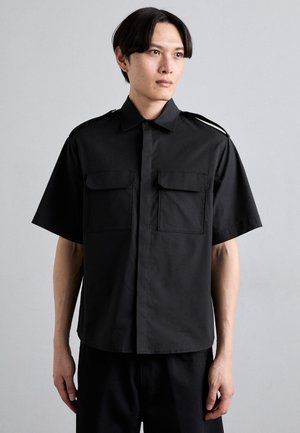 Camisa negra de manga corta hecha de tela suave, con dos bolsillos frontales, cuello abotonado y detalles en los hombros para un look estructurado.