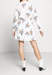 Mujer vestida con un vestido largo de mangas largas blanco con flores y cintura ceñida, usando botas de tobillo blancas con tacón, de pie contra un fondo liso.