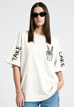 Femme portant un t-shirt blanc oversize avec un graphisme de main de paix et le texte "Take Care" sur les manches, associé à un jean bleu et des lunettes de soleil noires.