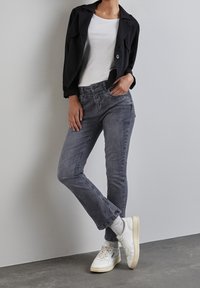 Persona con chaqueta negra corta, camiseta blanca, jeans grises, calcetines gris claro y zapatillas blancas, de pie contra una pared y suelo grises.