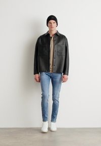 Camisa de cuero negra con dos bolsillos frontales, llevada sobre una blusa beige, combinada con jeans de color azul claro y zapatillas blancas, contra un fondo neutro.