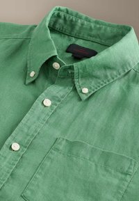 Chemise verte à boutons blancs, une poche avant et un col plié sur un fond beige.