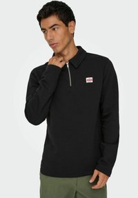 Pull noir avec un col zippé, des manches longues et une petite étiquette sur la poitrine. Fabriqué en tissu doux avec une texture uni.