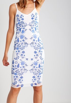 Robe blanche sans manches avec fines bretelles ornée de motifs floraux bleus symétriques, portée par une personne debout sur un fond uni.