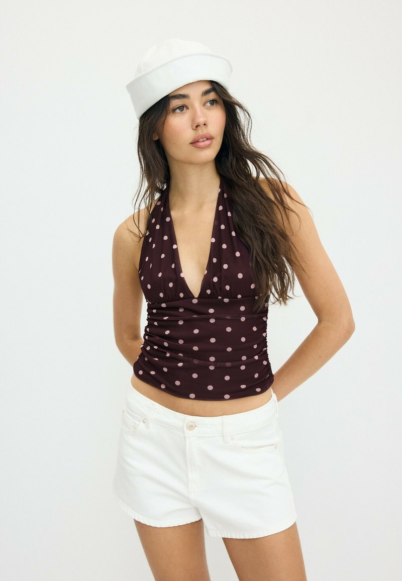 Jeune femme aux longs cheveux foncés portant un béret blanc, un haut marron à pois sans manches et un short blanc, debout devant un fond clair uni.
