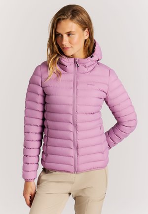 Frau trägt eine hellviolette gesteppte Jacke mit Kapuze, steht mit einer Hand in der Hüfte und schaut leicht zur Seite.