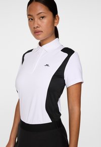 Jonge vrouw met donker haar, gekleed in een wit en zwart poloshirt met korte mouwen en zwarte onderkleding, die recht vooruit kijkt met een neutrale uitdrukking.