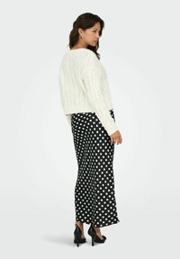 Maglione bianco a treccia abbinato a pantaloni a gamba larga neri con pois, che mostrano una superficie testurizzata e una vestibilità rilassata. Tacchi neri completano il look.