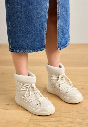 Pieds portant des baskets montantes beiges avec des lacets épais, associées à une jupe en jean bleue fendue à l'avant, debout sur un sol en bois.