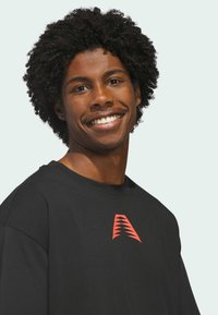 Black cotton t-shirt featuring a red geometric logo on the chest, round neckline, and relaxed fit, displayed on a smiling male model.