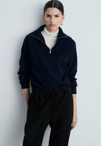 Námornícky zip-up mikina s vysokým golierom a dlhými rukávmi, skombinovaná s čiernymi elastickými nohavicami so širokým pásom. Hladká textúra, voľný strih.