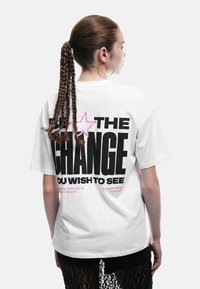 Ung kvinde med fletponytail iført hoopsøreringe og hvid t-shirt med stor sort og pink tekst på ryggen.