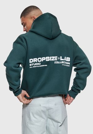 Mężczyzna w ciemnozielonej bluzie z kapturem z białym napisem "DROPSIZE LAB COLLECTION" na plecach, ubrany w jasne spodnie, stoi na białym tle.