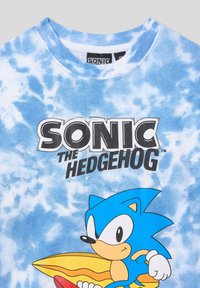 T-shirt blu tie-dye con una grafica di Sonic the Hedgehog in vivaci tonalità di blu e giallo, accompagnata da un testo nero in grassetto sopra. Materiale in cotone morbido.