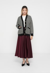 Black and white houndstooth cardigan with gold buttons, worn over a white top and maroon skirt, featuring long sleeves and pockets.