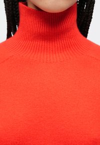 Czerwony sweter z golfem wykonany z miękkiej dzianiny. Posiada prążkowany kołnierz i gładką teksturę, prezentując jasny, jednolity kolor.