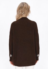 Cardigan călduros din tricot cu o croială lejeră, având un guler înalt și accente brodate albe pe mâneci. Textură moale.
