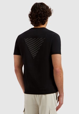 Man met krullend haar, gekleed in een zwart T-shirt met een donker gestreept omgekeerd driehoekontwerp op de rug en lichte cargobroek, staand met de rug naar de kijker.