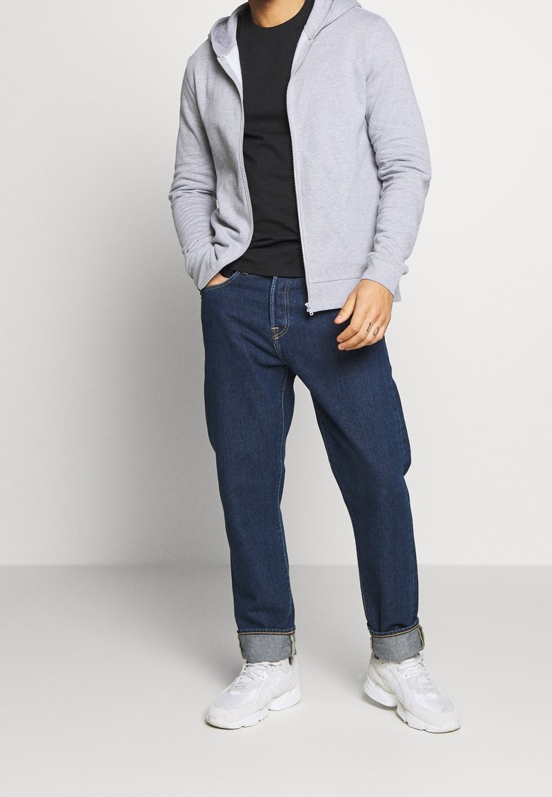 Grå hættejakke med lynlås over en sort skjorte, parret med mørkeblå jeans og hvide sneakers; afslappet, komfortabel pasform, glat tekstur.