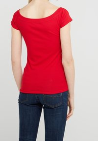 Röd kortärmad topp med off-the-shoulder halsringning, tillverkad av mjukt material, kombinerad med blå denimjeans med bakfickor med sömmar.