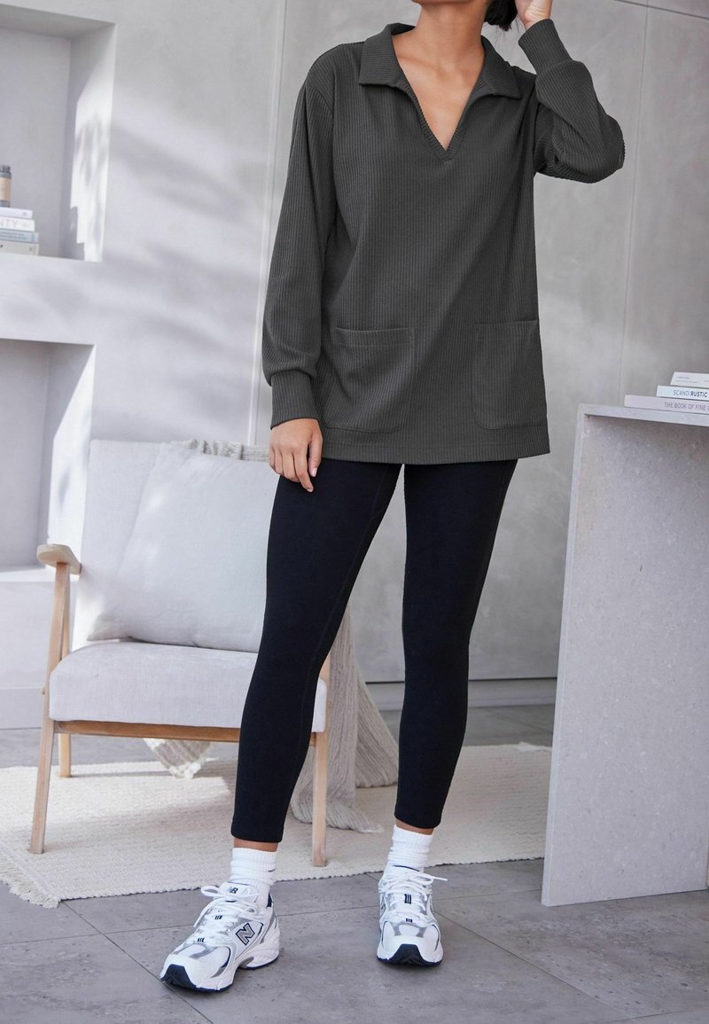 Haut à manches longues côtelé gris foncé avec un décolleté en V, deux poches avant, associé à des leggings noirs et des chaussures de sport blanches avec des accents argentés.