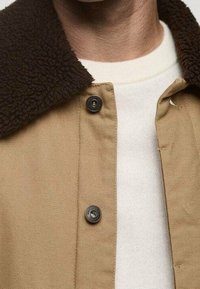 Beżowa kurtka z brązowym, teksturowanym kołnierzem z polaru, z czarnymi zapięciami na zatrzaski i kremowym swetrem pod spodem.