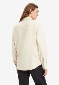 Camisa de manga larga color beige claro, hecha de una tela suave, con cuello puntiagudo, puños con botón y un detalle de yugo en la parte posterior.