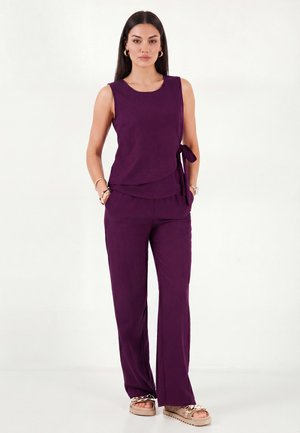 Woman with long dark hair wearing sleeveless purple top with side tie and matching pants, standing with hands in pockets and neutral expression.