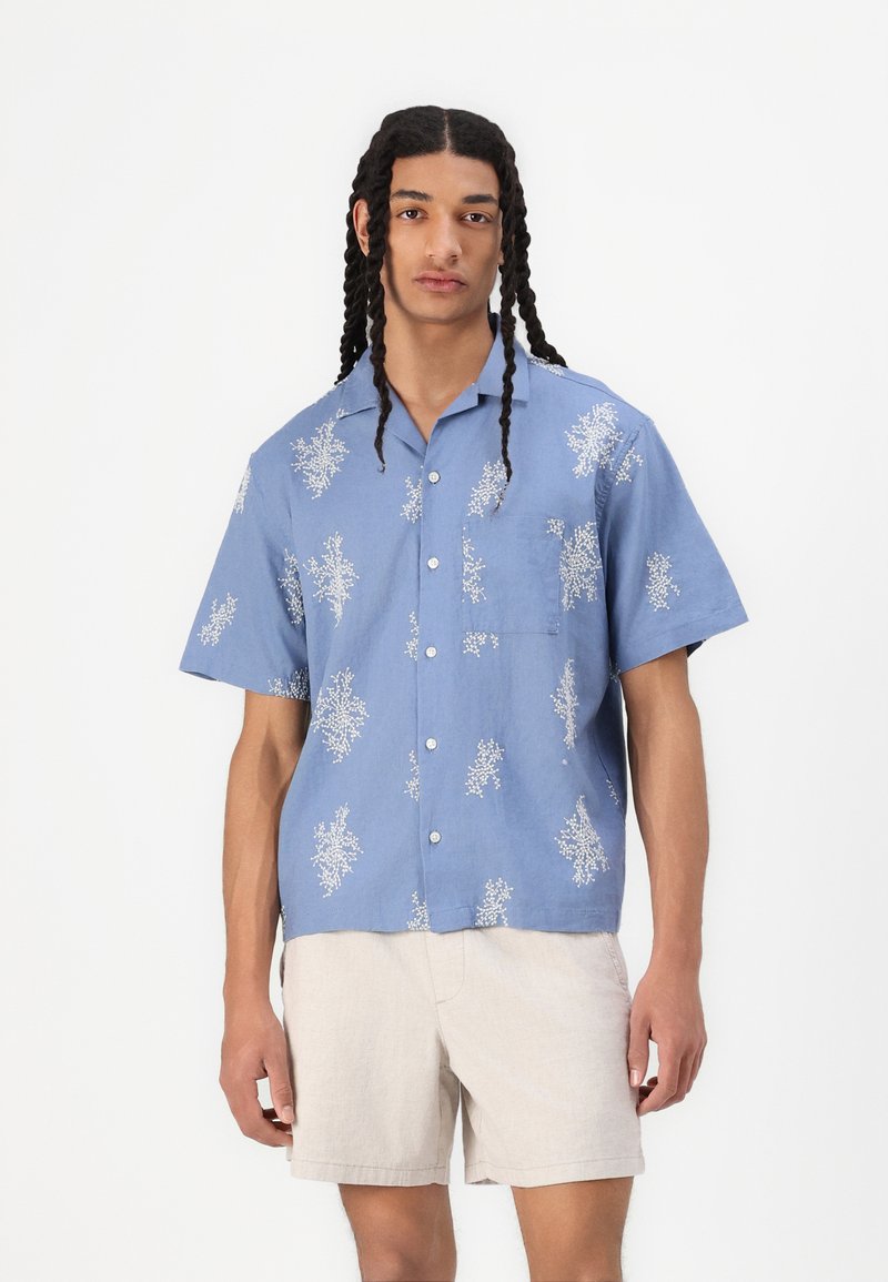 Chemise bleue à manches courtes avec des motifs floraux blancs, devant boutonné et poche poitrine ; associée à un short beige clair. Texture de tissu douce.