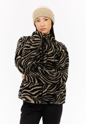 Jonge vrouw die een beige gebreide beanie en een zwart-gele zebra-gestreepte fuzzy jas draagt, met zwarte broek, terwijl ze met haar hand bij haar gezicht poseert.