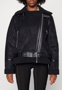 Chaqueta bomber de ante negro con cuello de piel de oveja, detalles de cremallera y hebilla, y cintura con cinturón. Presenta bolsillos con cremallera y puños texturizados.