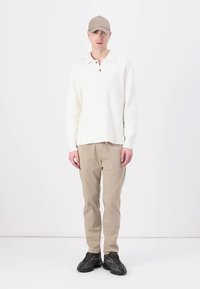 Valkoinen fleecepaita pitkähihaisella kauluksella, beige chinopuvut ja mustat kuviolliset lenkkarit. Mallilla myös beiget lippalakki.