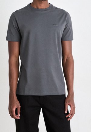 Muž v jednobarevném šedém tričku s krátkým rukávem a logem "BOSS" na hrudi, v černých kalhotách, stojící před bílým pozadím.