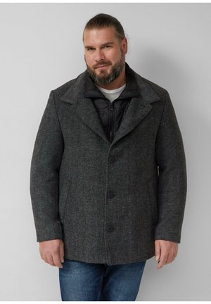 Bärtiger Mann mit zurückgebundenem Haar, der einen dunkelgrauen, geknöpften Mantel über einer schwarzen Zip-up-Jacke und blauen Jeans vor einem schlichten Hintergrund trägt.