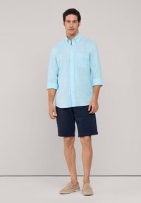 Camisa de rayas azul claro con cuello abotonado, bolsillo en el pecho y mangas enrolladas, combinada con pantalones cortos azul marino oscuro y zapatos de lona beige. 