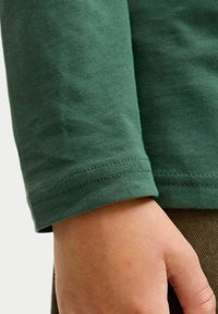 Grøn langærmet skjorte lavet af bomuld, med en glat tekstur, tætsiddende manchetter og et enkelt design. En hånd er delvist synlig, hvilende på stoffet.